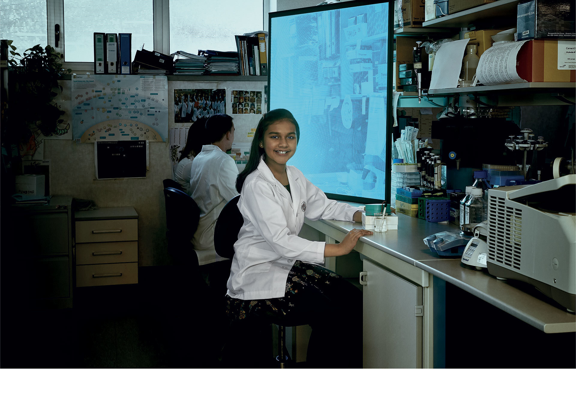 Two researchers in white lab coats work in a laboratory, one smiling at camera, with equipment and a large screen in background.