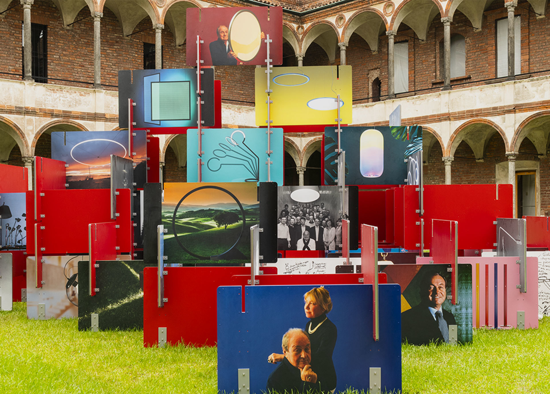 Colorful outdoor installation with red panels, photos, and Artemide lamp images in a historic arcaded courtyard.