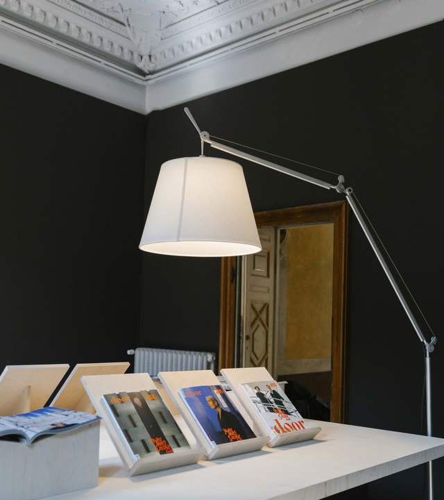 Tolomeo floor lamp with illuminated shade lighting a display table with door magazines, in an interior with dark walls and a decorated ceiling.