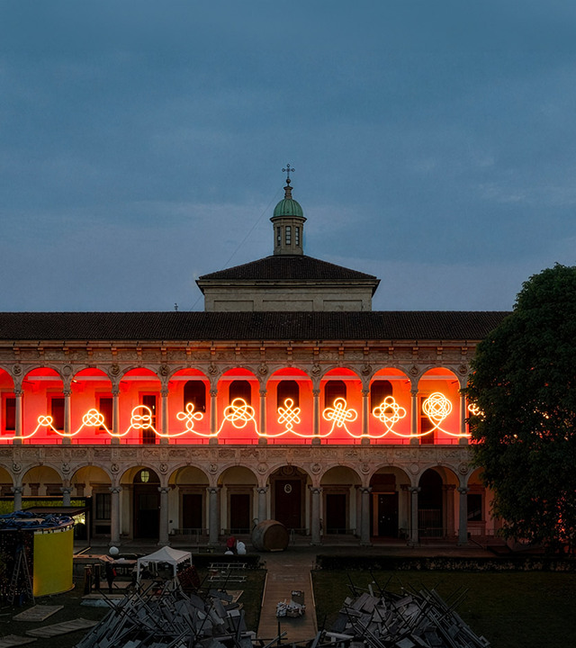 Facade of the courtyard of the University of Milan at dusk, with a linear light installation drawing interwoven motifs across the arches.