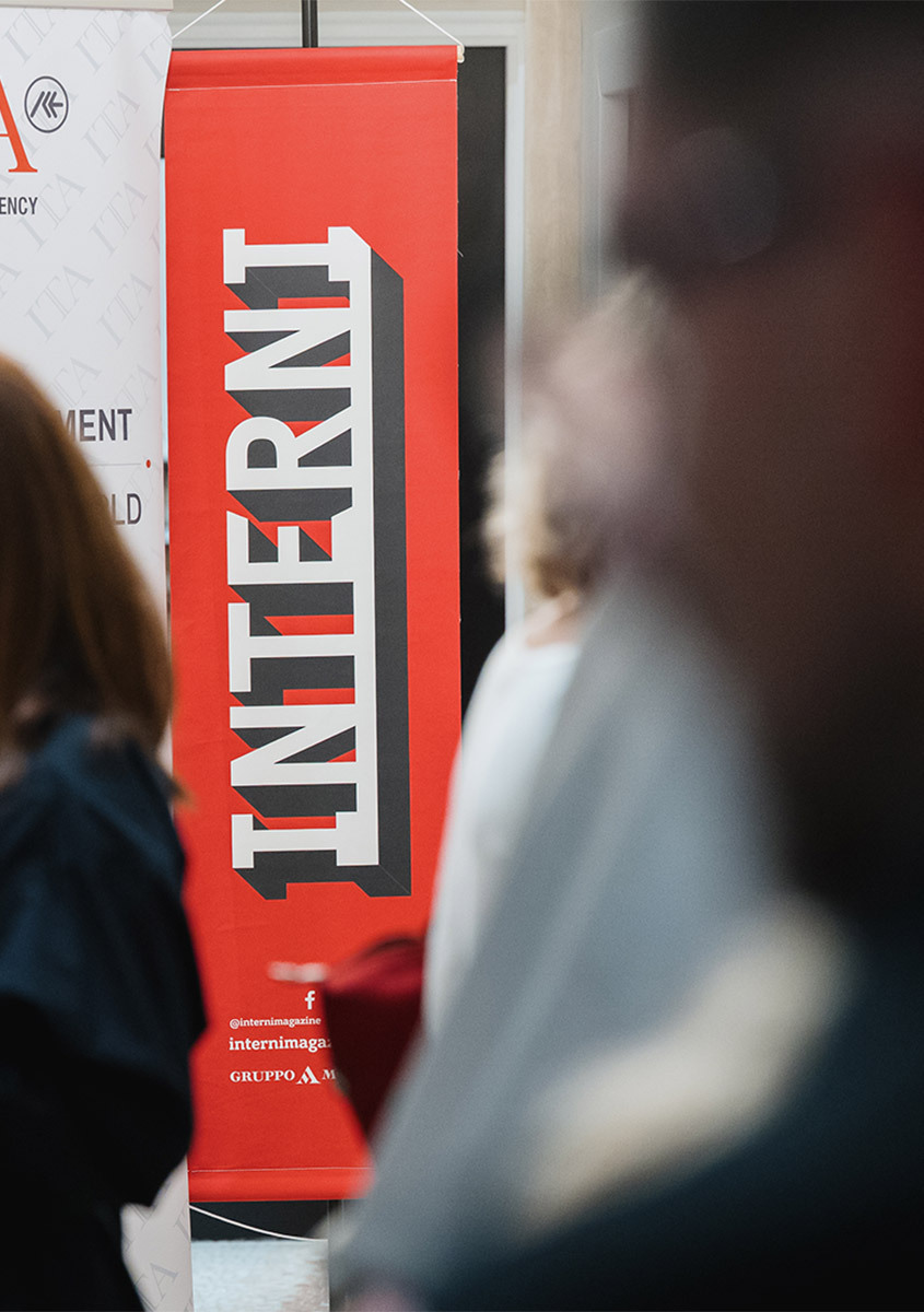 Red vertical banner with bold 'INTERNI' text displayed at a design event, with blurred attendees in the foreground.