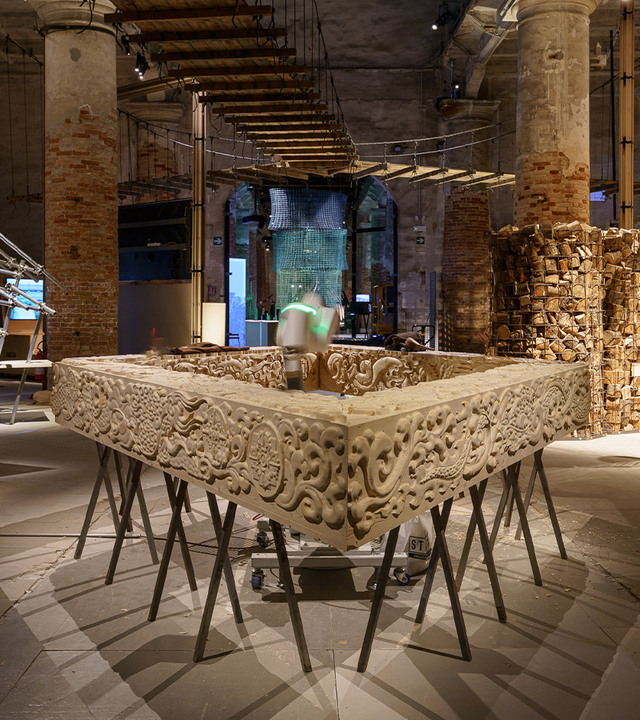 Art installation with a robot sculpting a diamond-shaped wooden sculpture.
