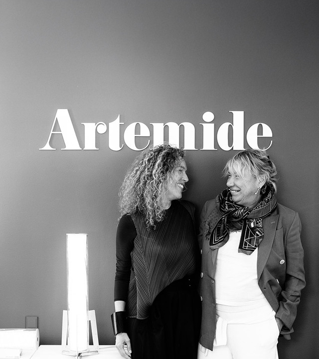 Black and white portrait of Carlotta de Bevilacqua and Giulia Foscari standing beneath the Artemide logo, next to the Criosfera lamp.