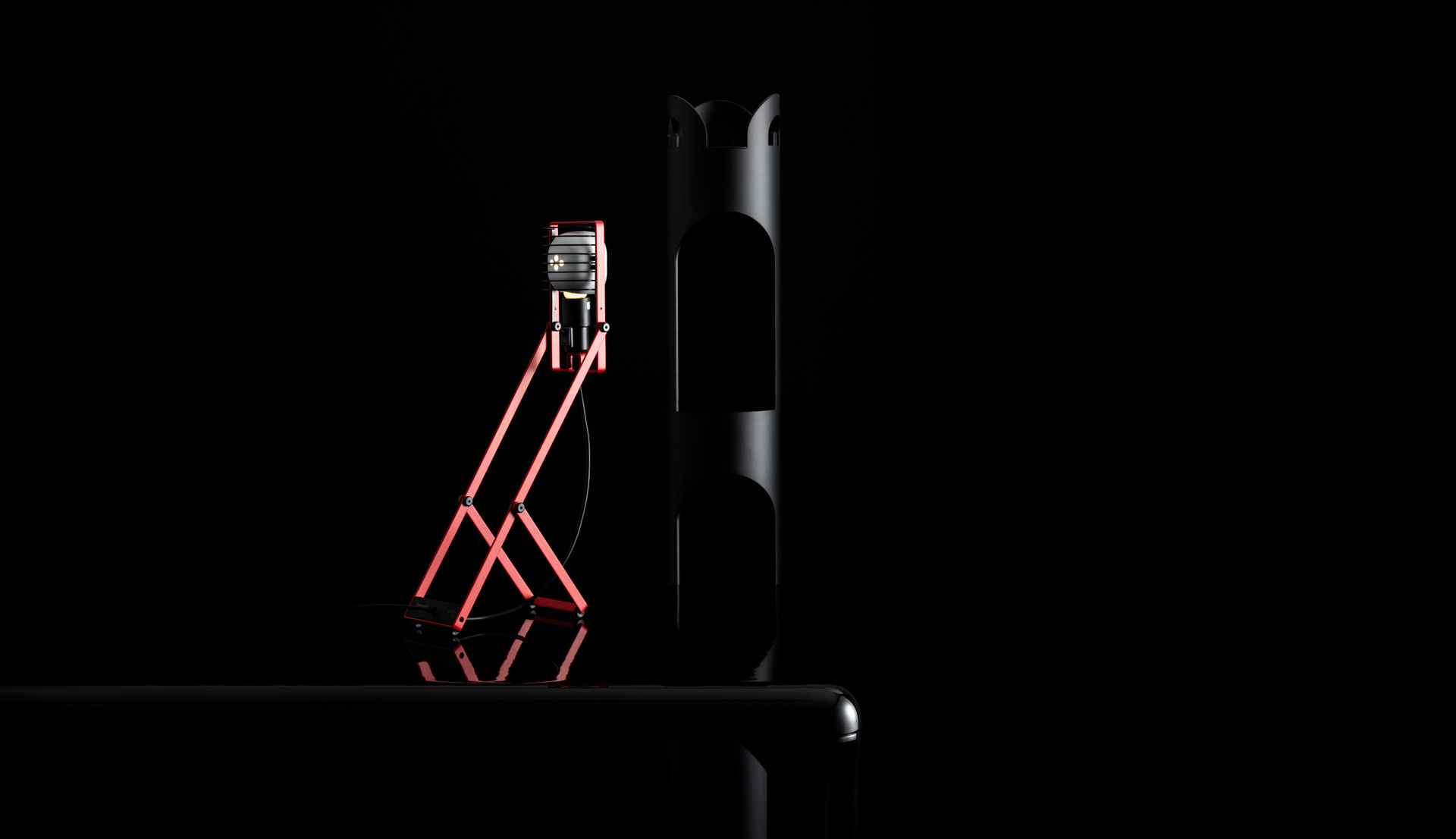 Red Sintesi lamp placed on a table, next to a black coat rack, against a dark background.
