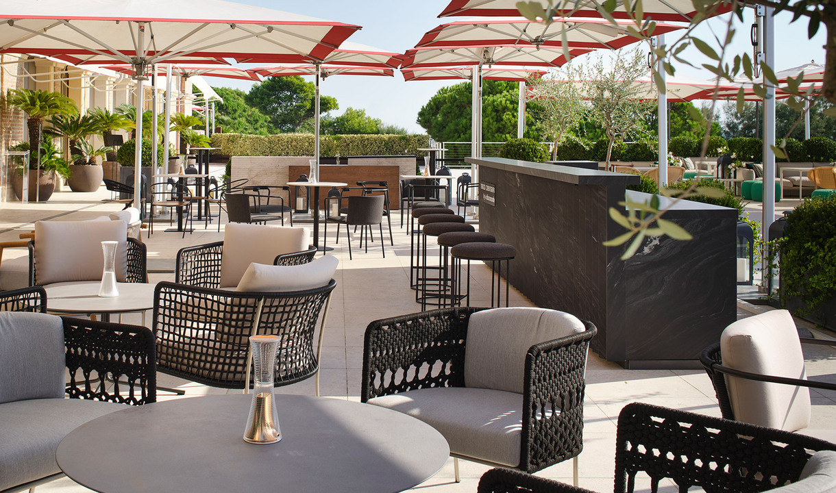 Elegant outdoor terrace with woven black lounge chairs, small round tables with a slim table lamp, large umbrellas, and a bar counter.
