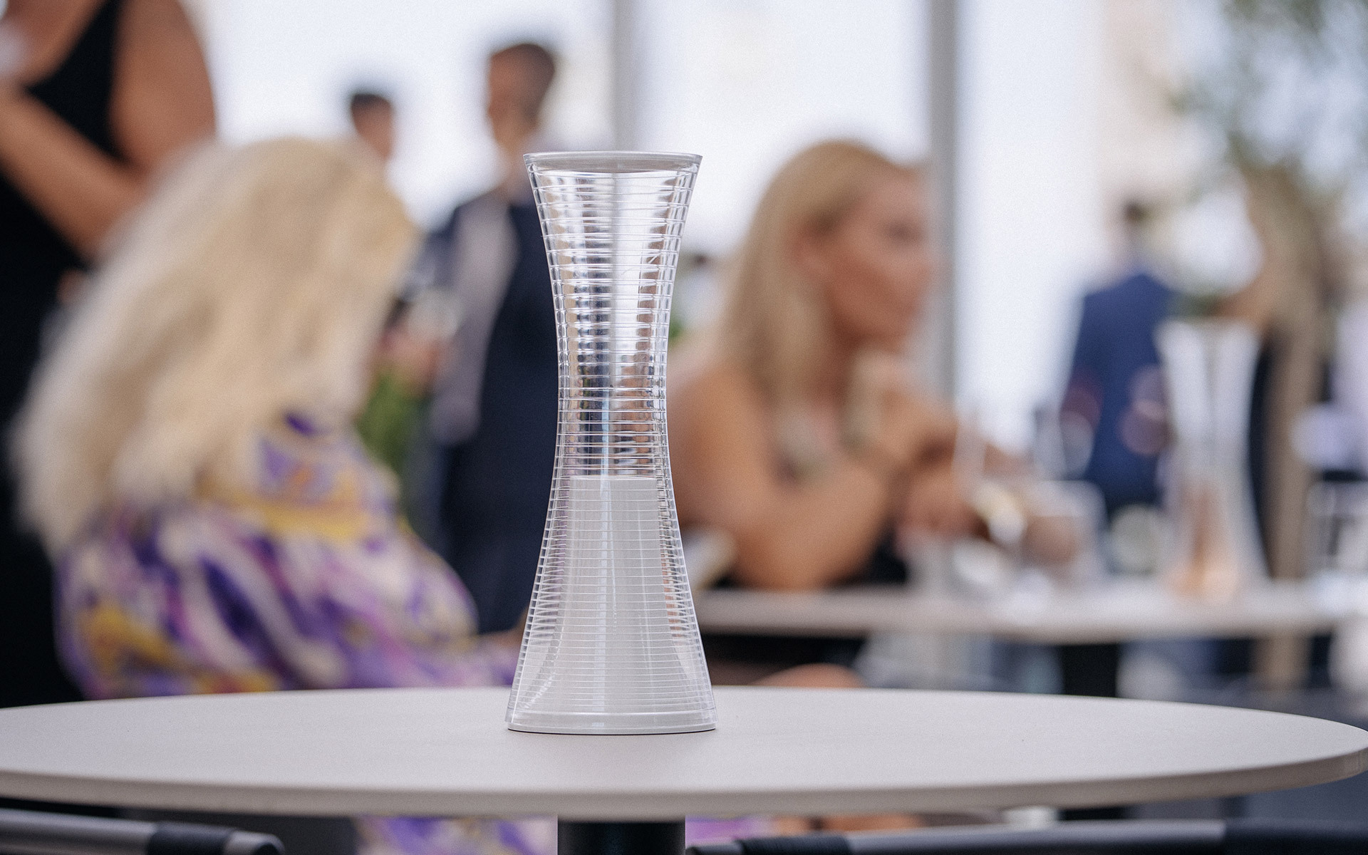 artemifgitt.sbse Together portable table lamp, hourglass-shaped transparent diffuser, displayed on a white table at an elegant social event