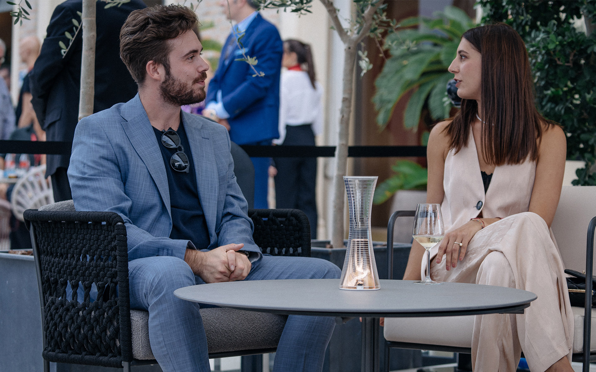 Two people conversing at an outdoor café table with an Artemide glass candle lantern centerpiece and a wine glass.