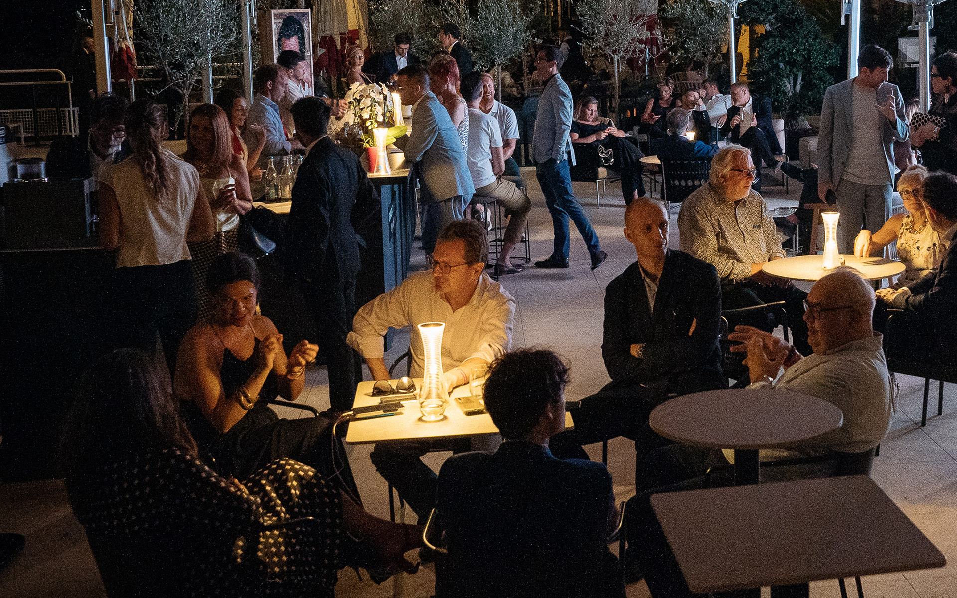 Outdoor evening gathering with people socializing at tables, illuminated by a warm glowing portable table lamp centerpiece.