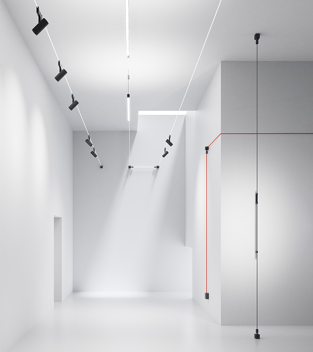 Artemide cable lighting system in a white minimalist space: black adjustable spotlights on tensioned wires illuminate walls and floor.