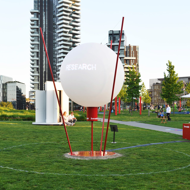 Artemide large white spherical outdoor installation labeled 'Research' on red metal poles in an urban park with modern buildings.
