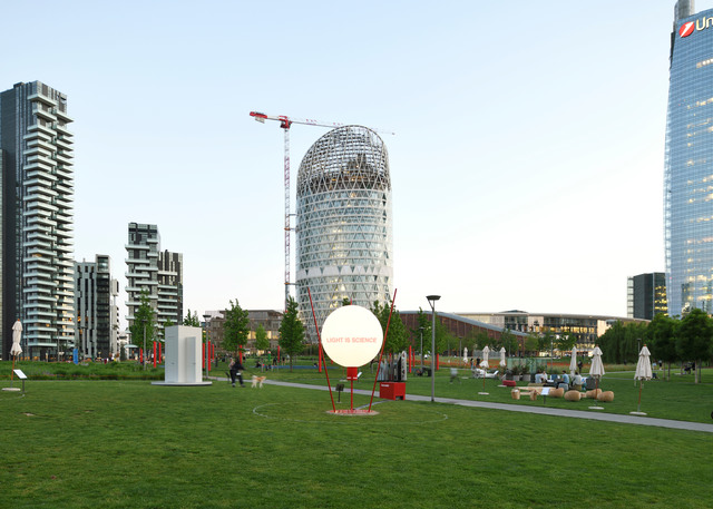 Artemide spherical outdoor lamp on red stand in Milan's Porta Nuova park, with modern skyscrapers and green lawn at dusk