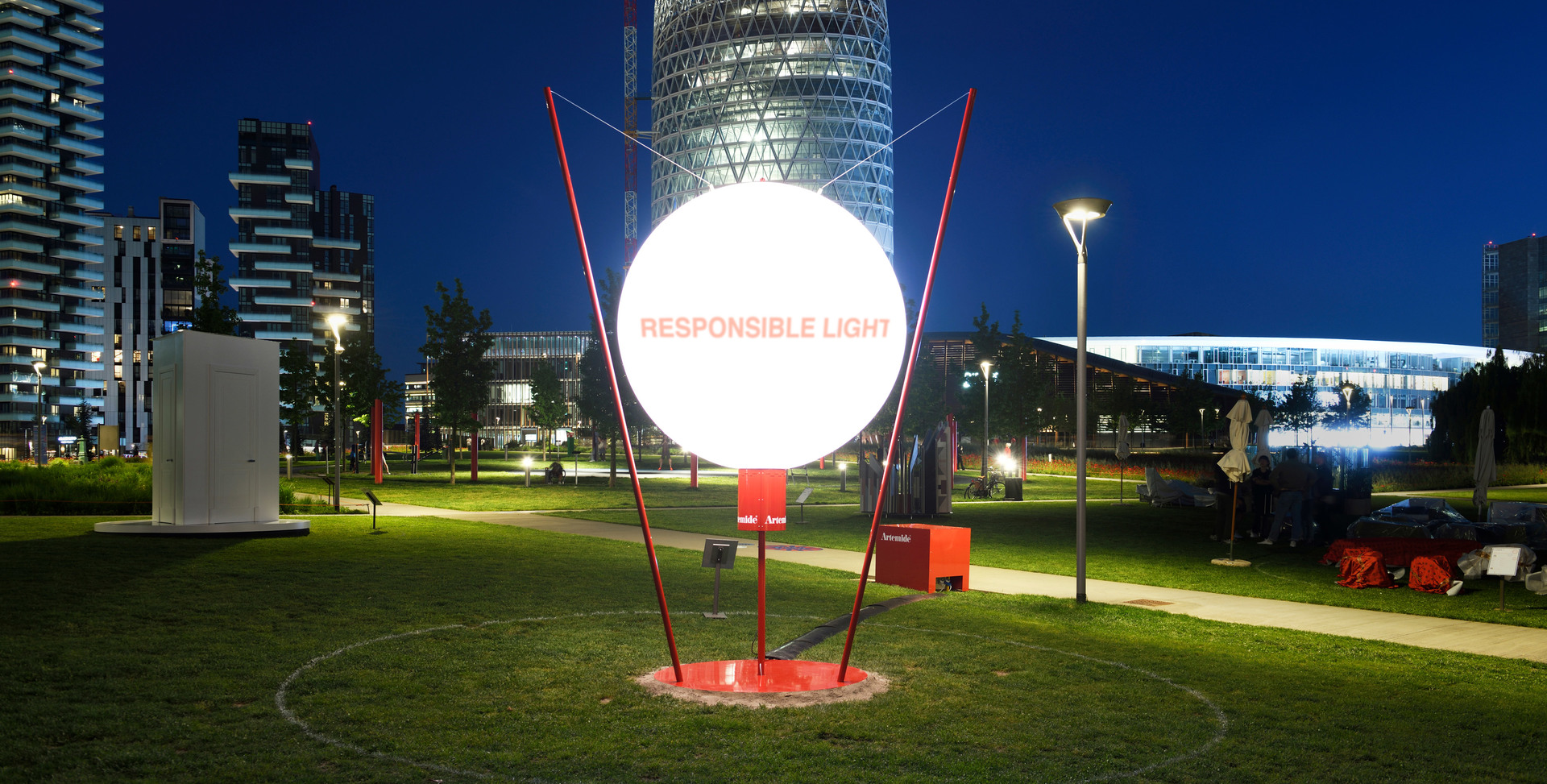 Artemide 'Responsible Light' installation: glowing white sphere on red metal structure in urban park at night, city skyline behind