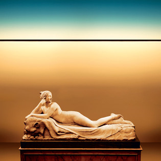 Reclining classical statue on a pedestal, lit by Dusk, a thin horizontal light line, in a warm-toned space with gradient lighting. Photo by Pierpaolo Ferrari.