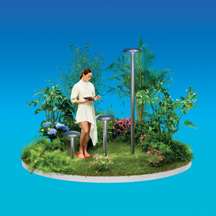 Woman standing on a small green island with plants and flowers beside Knop outdoor lamps against a blue background. Photo by Pierpaolo Ferrari.
