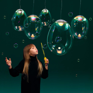 Child holding a bubble wand beneath a group of Stellar Nebula lamps, surrounded by bubbles against a dark green background. Photo by Pierpaolo Ferrari.