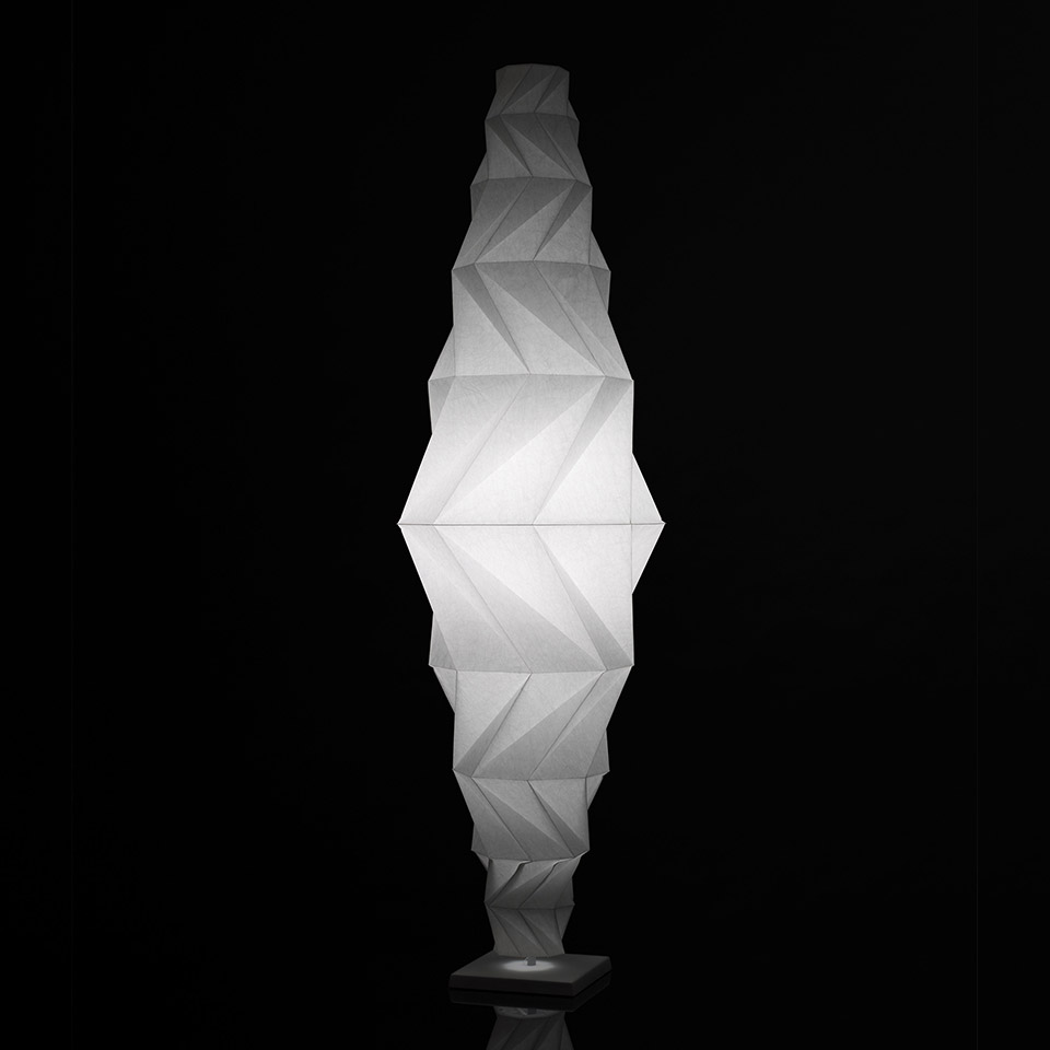 Artemide In-Ei Issey Miyake Minomushi floor lamp: tall origami-folded white fabric shade, diamond-shaped, glowing softly on black base