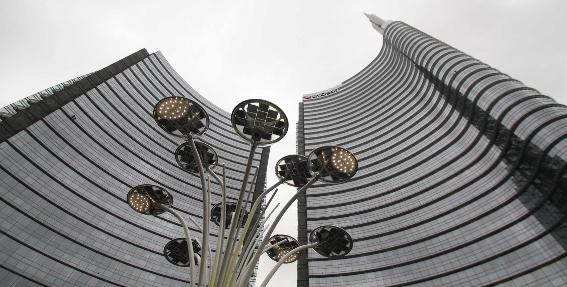 Artemide outdoor LED street lamps with circular disc heads on curved stems, set against the curved glass facade of UniCredit Tower, Milan