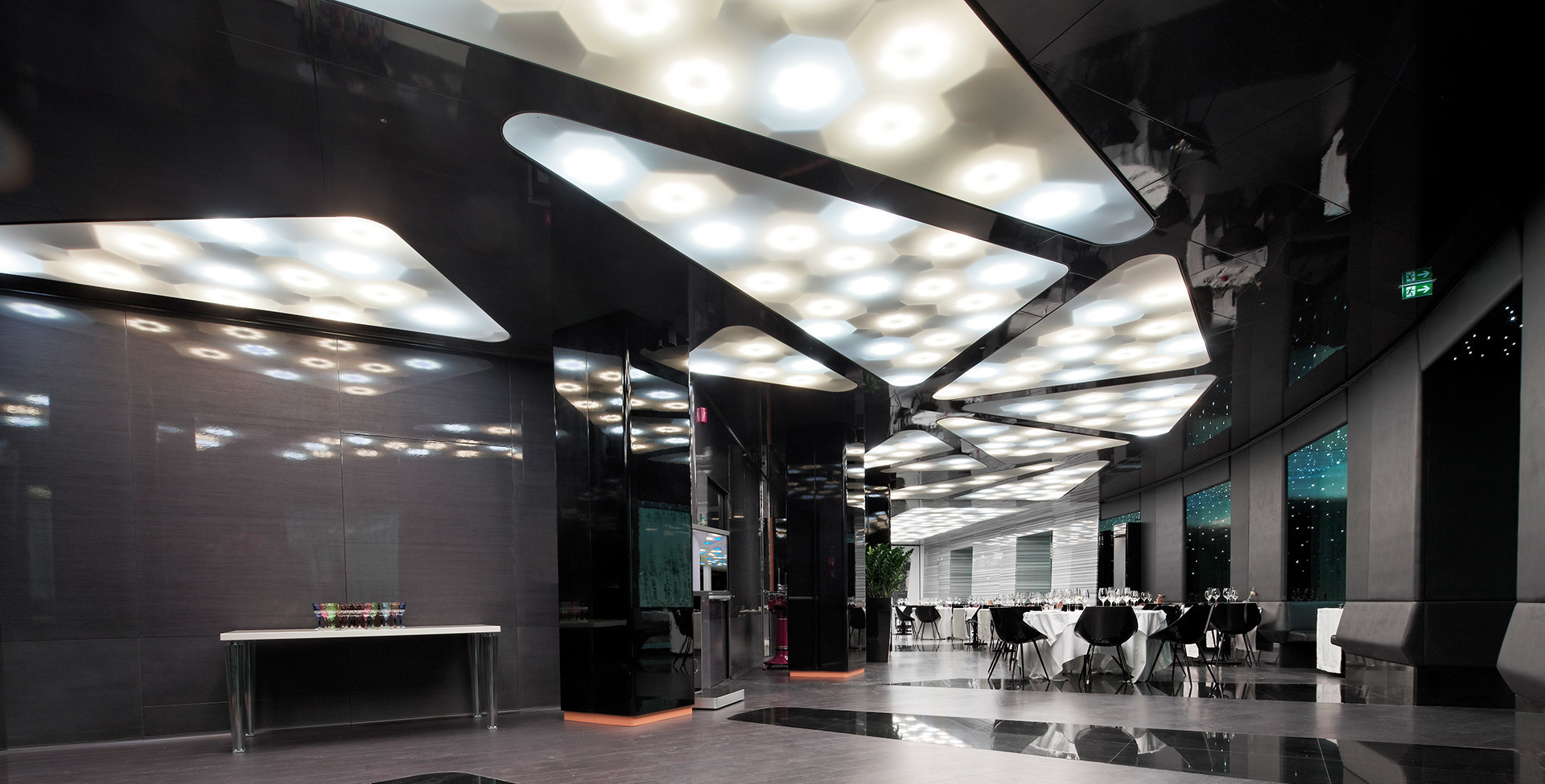 Modern interior with honeycomb-patterned ceiling lights illuminating a sleek dark lobby with dining area in the background