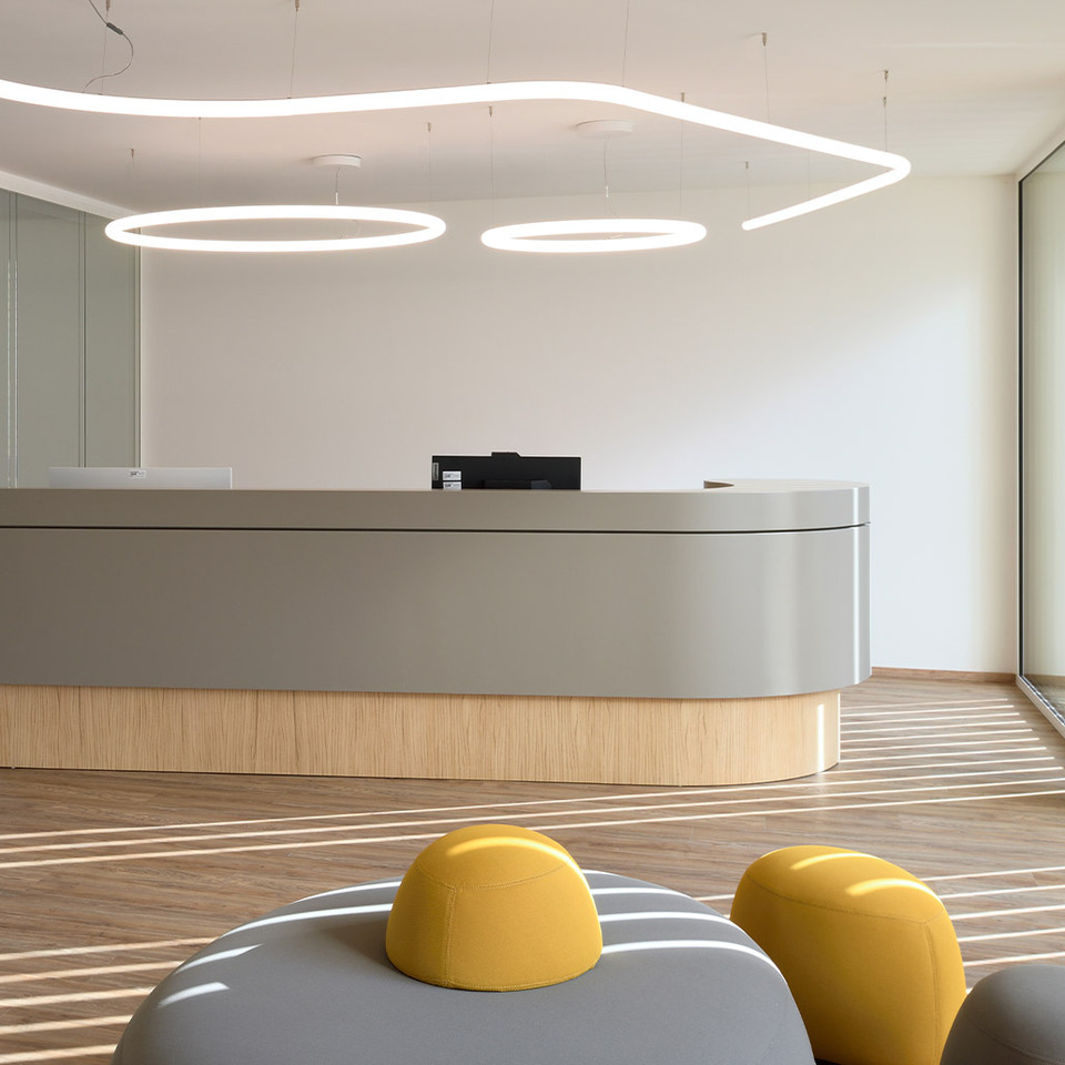 Reception area of the new Fornaroli Polymers headquarters with a curved desk, wooden flooring and glass walls, illuminated by the Alphabet of Light suspension system.