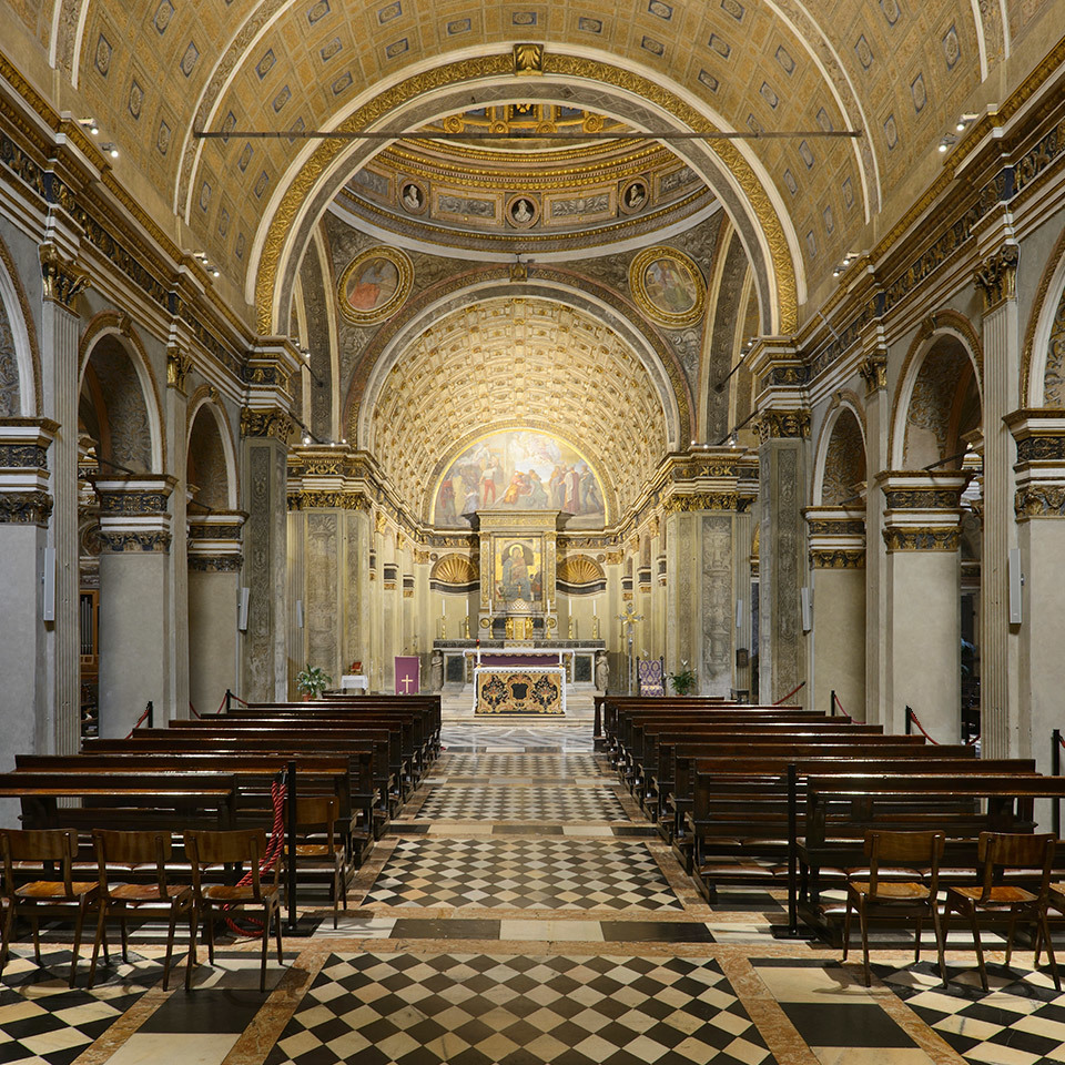 Interior of a Renaissance church with gilded vaulted ceilings, frescoed apse, marble altar, checkered floor, and wooden pews in two rows.