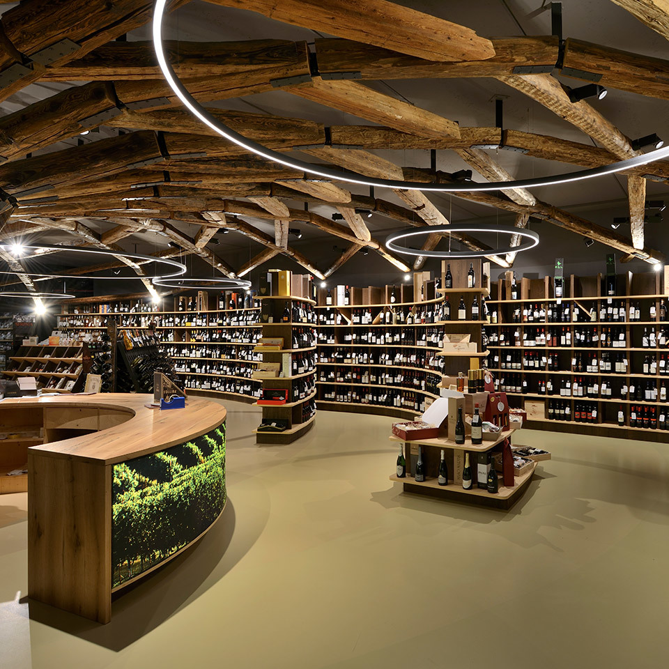 Wine shop interior with Artemide circular pendant lights among rustic wooden ceiling beams, illuminating curved shelves stocked with bottles.