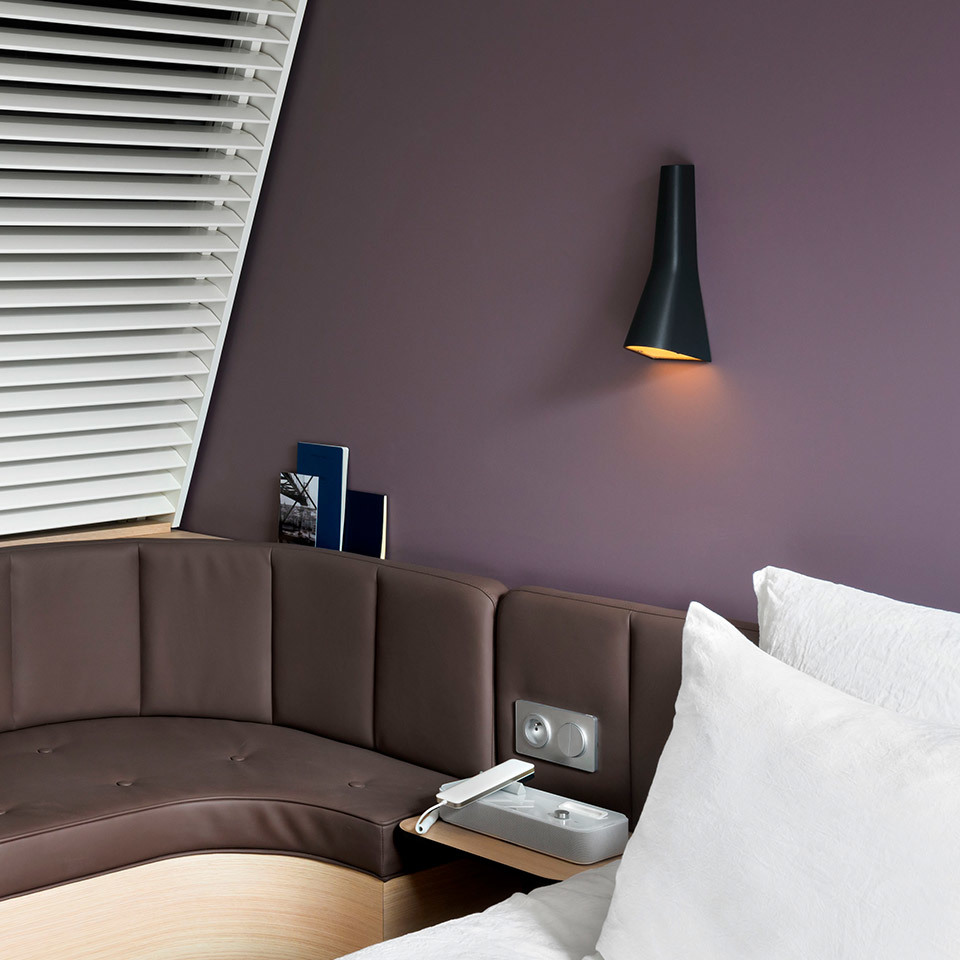 Black conical Artemide wall lamp with gold interior mounted above a hotel bed headboard on a deep purple wall.