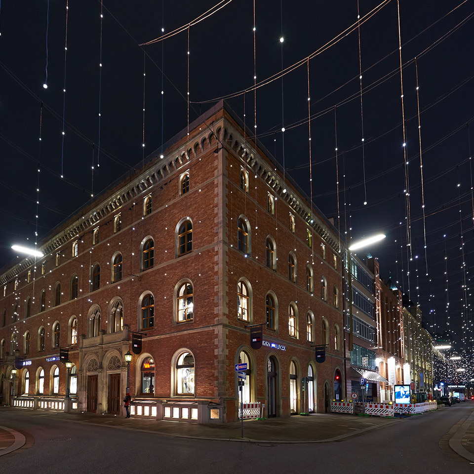 Historic red-brick corner building illuminated at night with linear LED street lights and decorative string lights overhead
