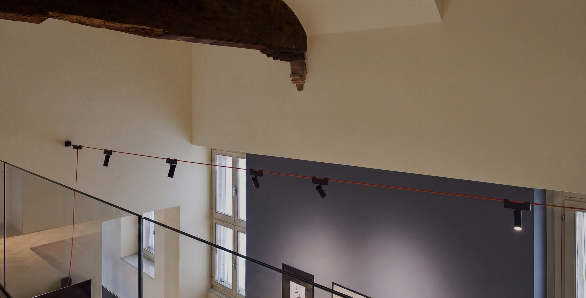 Funivia system with red cable and adjustable spotlights in a double-height space with historic wooden beams.
