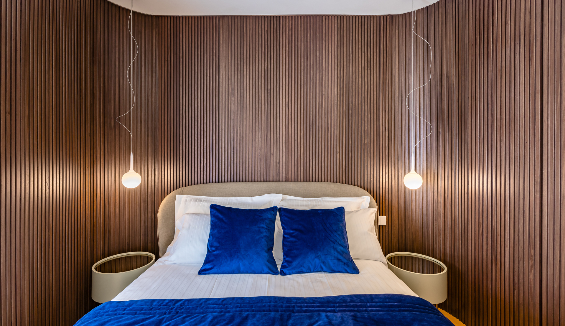 Bedroom with wood-paneled wall featuring two pendant lamps with spherical globes hanging on thin wires beside an upholstered bed with blue pillows
