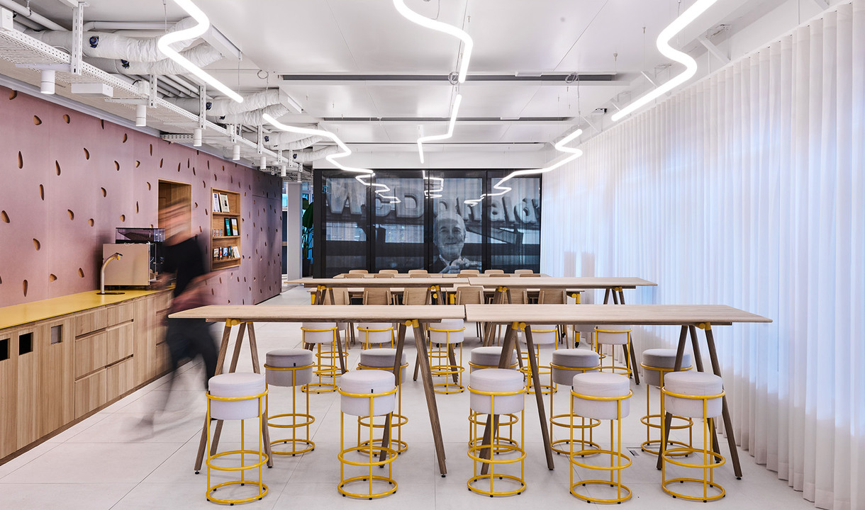 Corporate kitchen area with high tables, yellow stools and curved LED lights on the ceiling.
