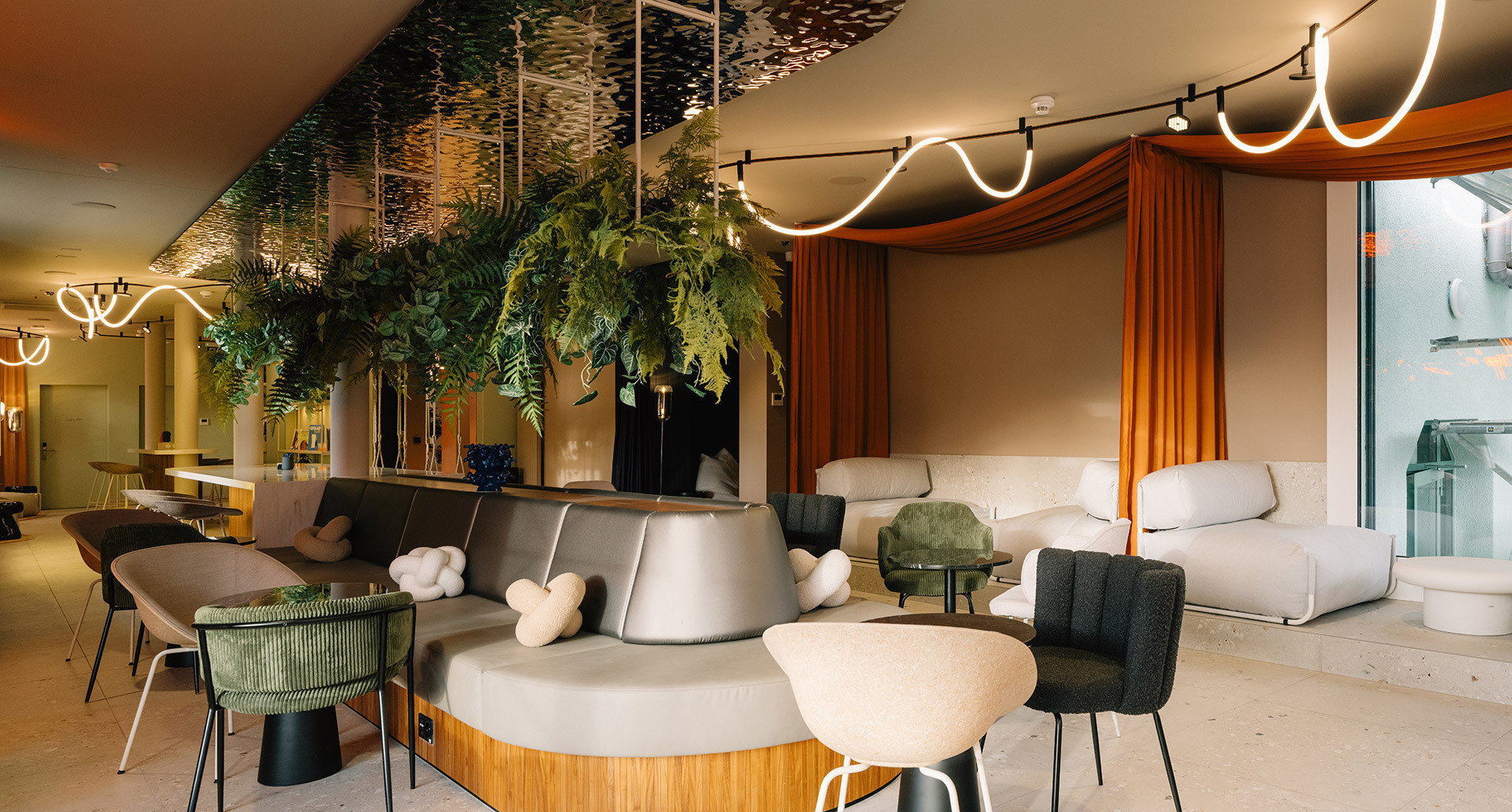 Spacious lounge with upholstered seating, hanging plants, orange curtains, and the Sylt ceiling system with luminous lines.