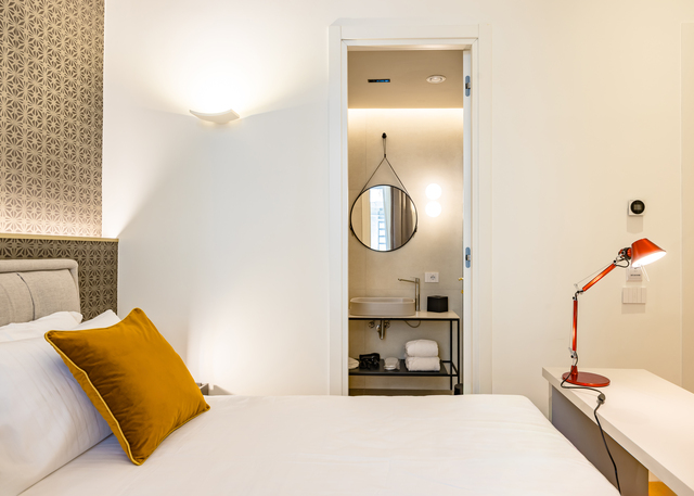 Hotel bedroom with red Artemide Tolomeo desk lamp on nightstand, wall sconce above headboard, and lit bathroom visible through doorway.