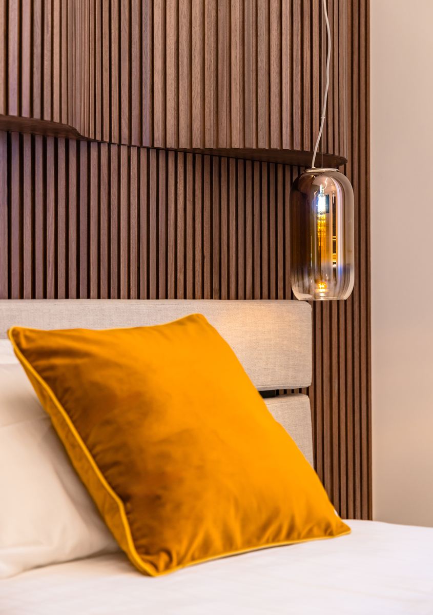 Artemide pendant lamp with clear glass capsule shade hanging beside a wooden slat headboard above a bed with mustard velvet pillow.