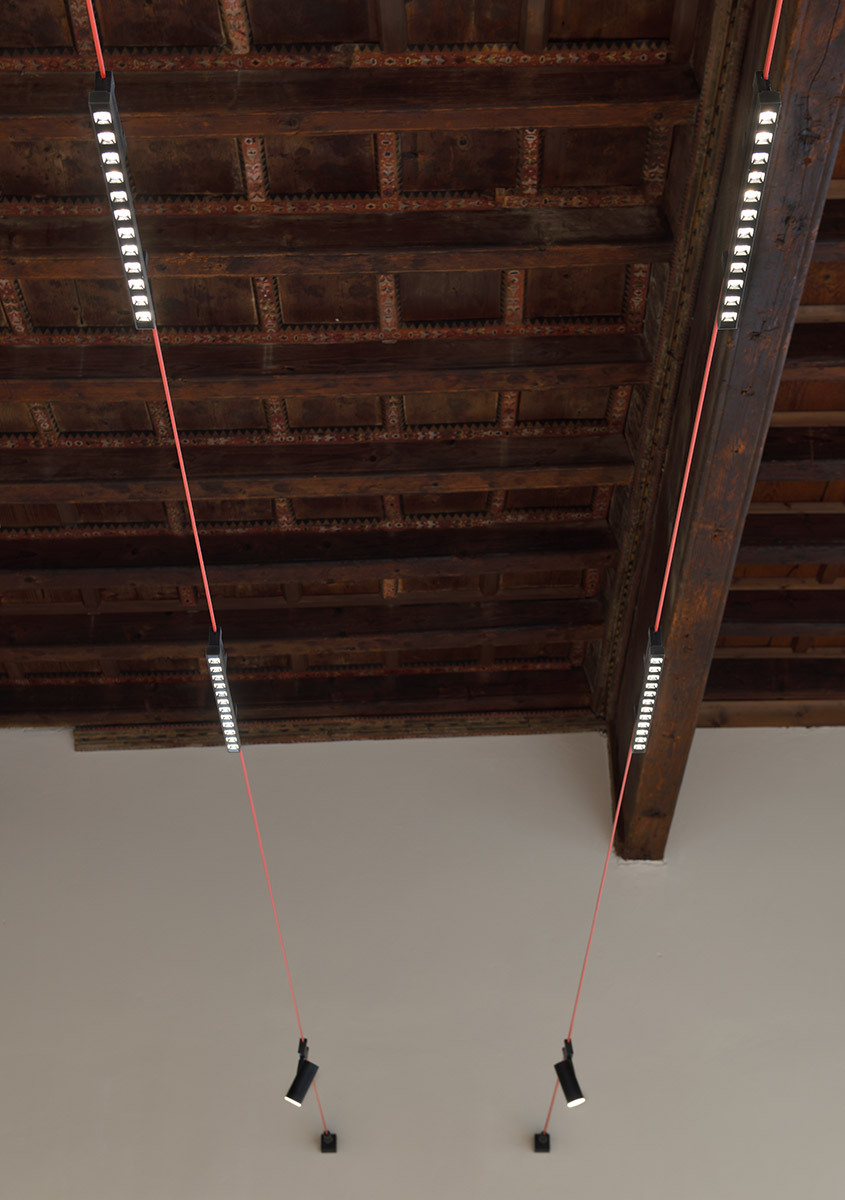 Funivia linear light modules and adjustable spotlights suspended beneath a historic decorated wooden ceiling.