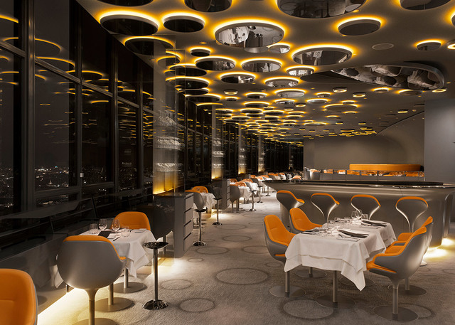 Elegant restaurant interior with dramatic ceiling of circular amber-lit Artemide fixtures, orange chairs, white-clothed tables, city view at night