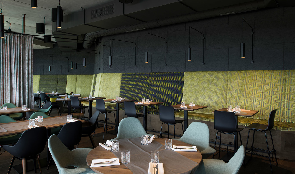 Restaurant interior with dark pendant cylinder lamps over wooden tables, teal chairs, and green upholstered wall panels.