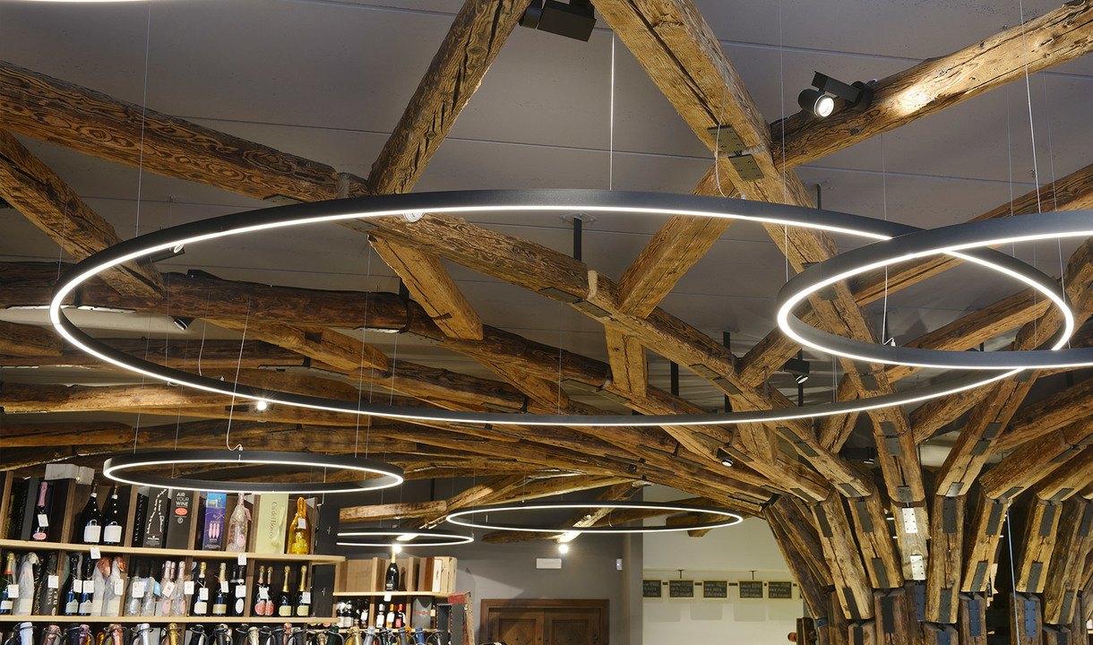 Multiple Artemide circular LED pendant rings of varying sizes suspended over a wine shop with rustic wooden beam ceiling.