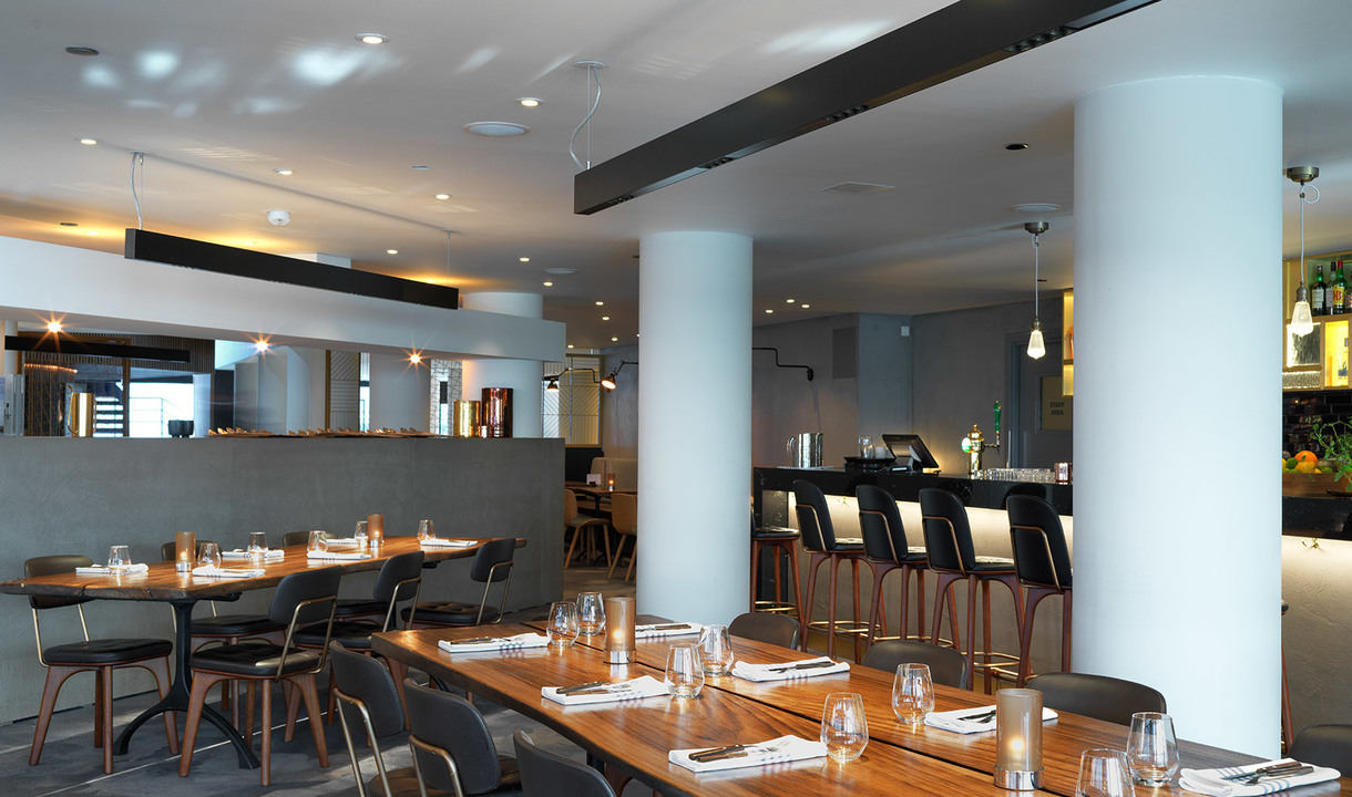 Modern restaurant interior with recessed ceiling lights, pendant lamps over wooden tables set with glassware, white columns, and a bar area.