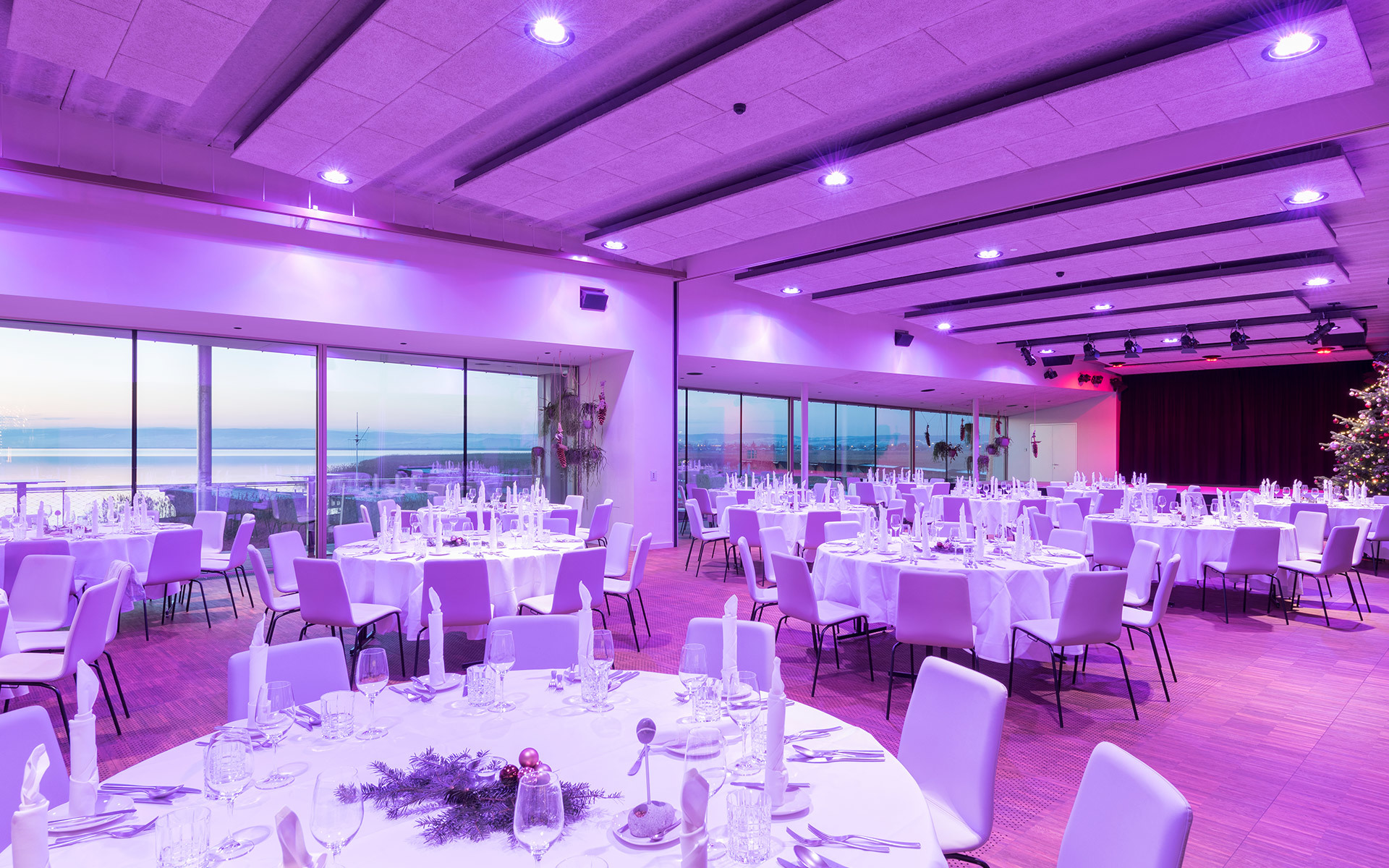 Elegant banquet hall with purple ambient lighting, round white-clothed tables set for dinner, and panoramic ocean view windows.