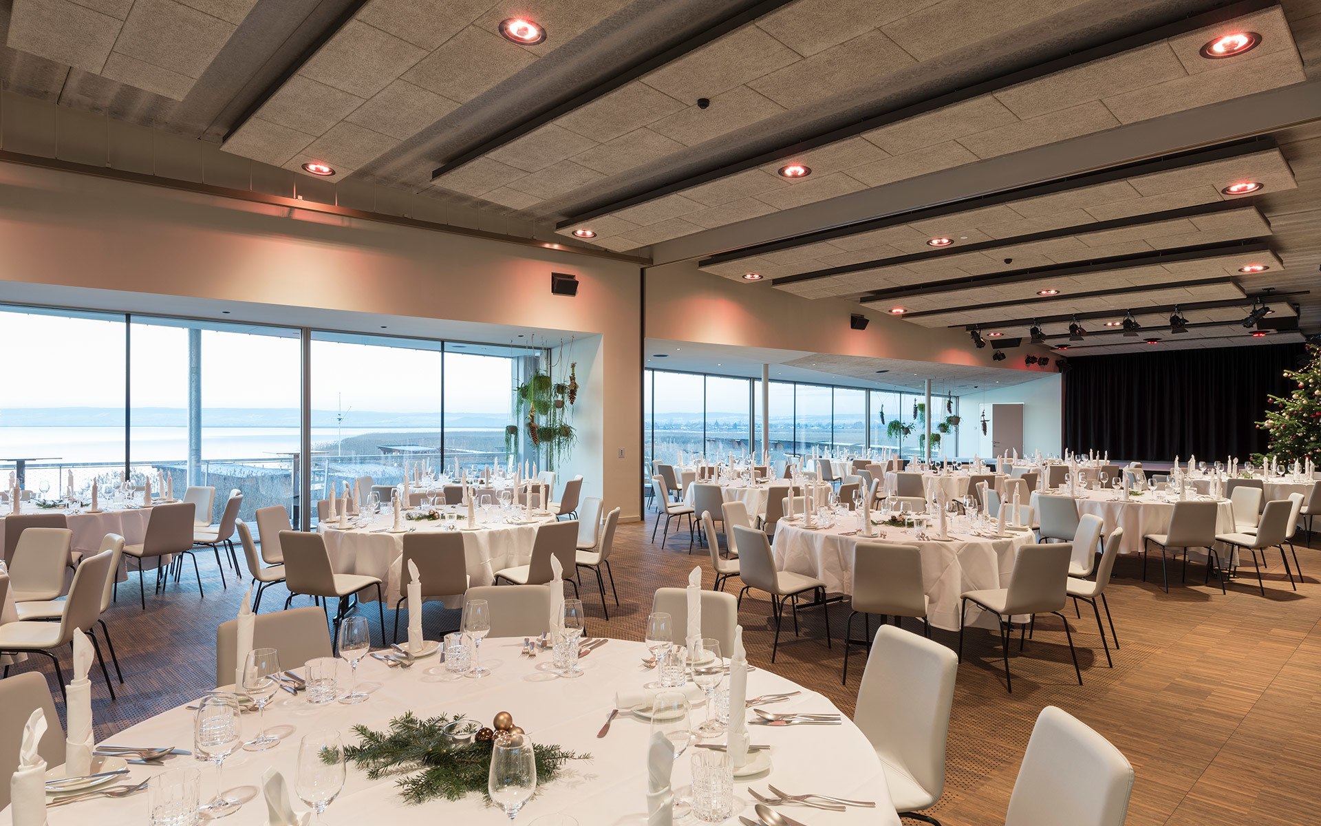 Elegant banquet hall with round white-clothed tables, recessed ceiling lights, and panoramic sea views through floor-to-ceiling windows.
