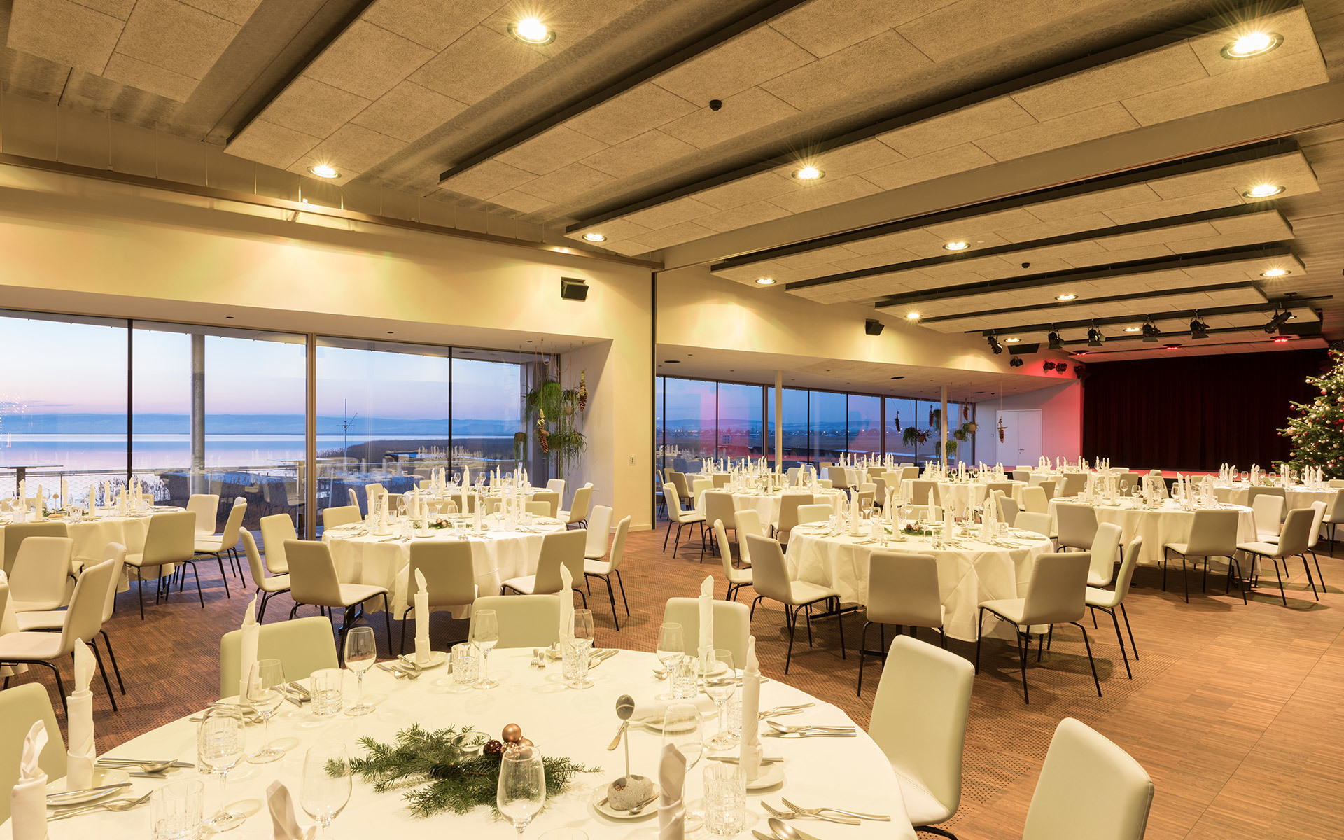 Elegant banquet hall with recessed ceiling spotlights, round white-clothed tables set for dinner, large windows with ocean sunset view.