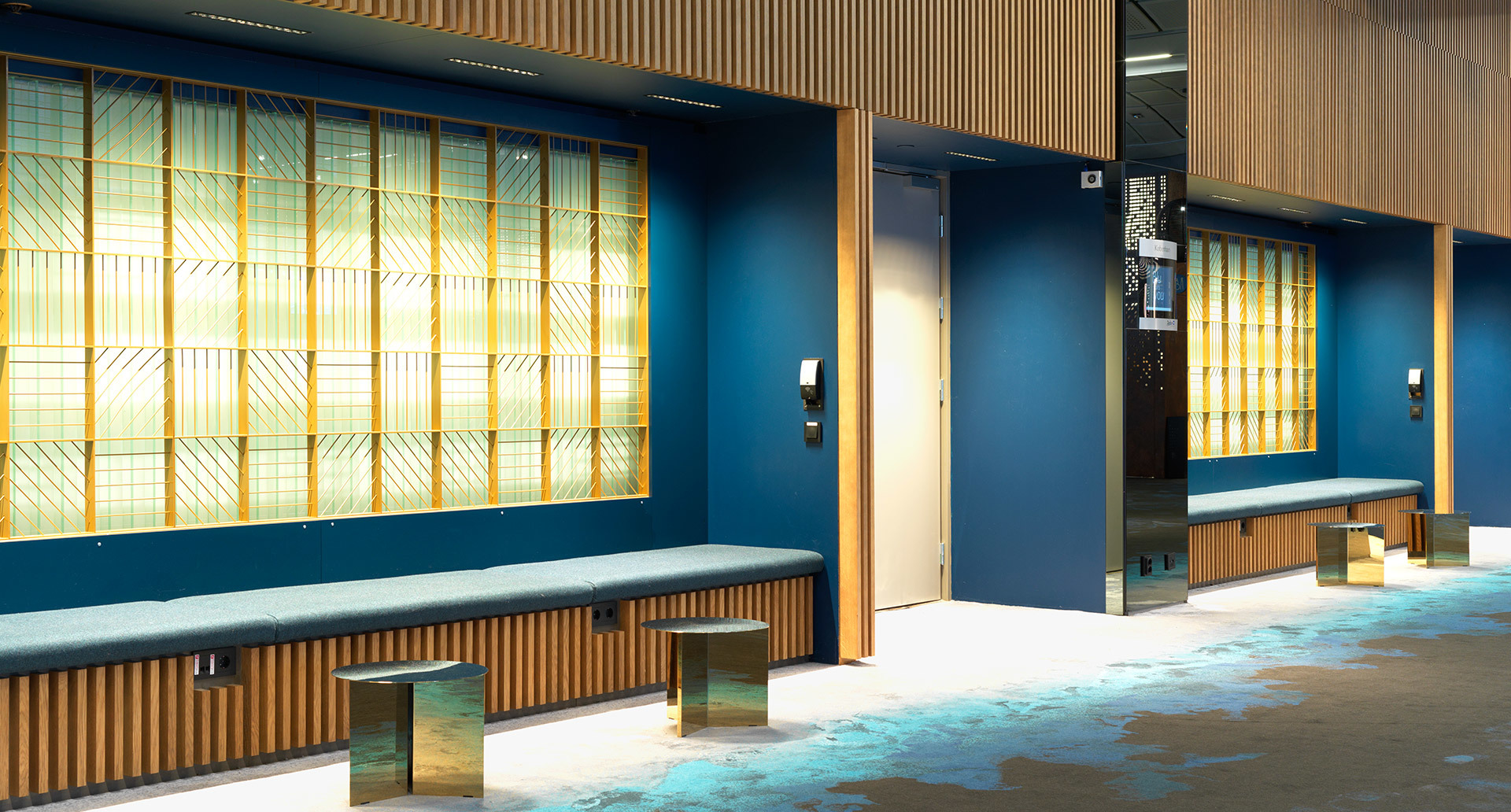 Modern lobby with teal walls, wood-paneled benches, glowing golden lattice backlit panels, and accent floor lighting on polished surfaces