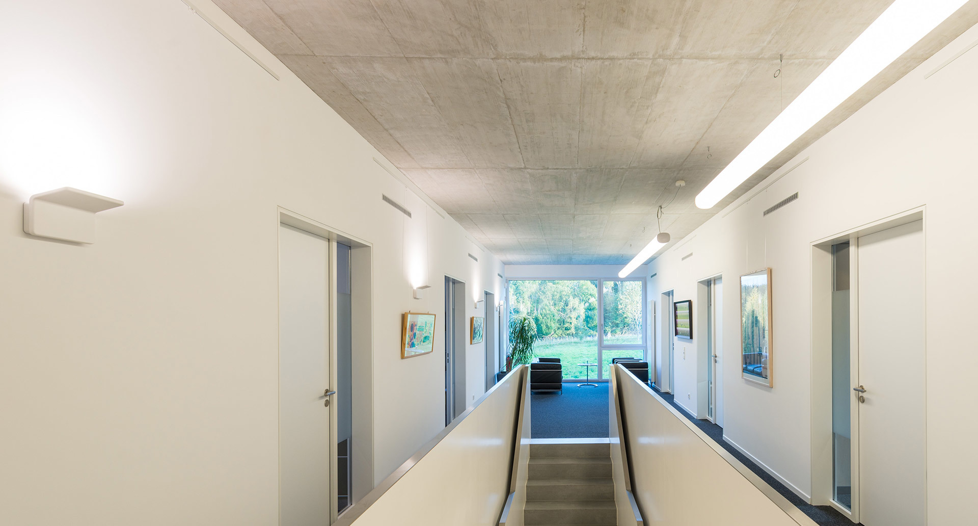 Modern corridor with white walls, concrete ceiling, linear ceiling lights, and wall-mounted uplights illuminating a long hallway with doors and artwork.