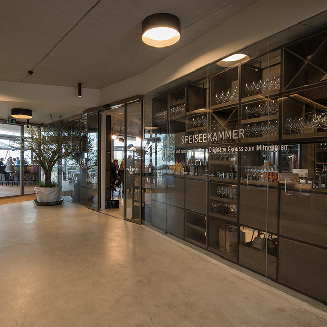 Modern corridor with Artemide ceiling light fixture illuminating a glass-walled gourmet shop 'Speiseekammer' displaying wine glasses.