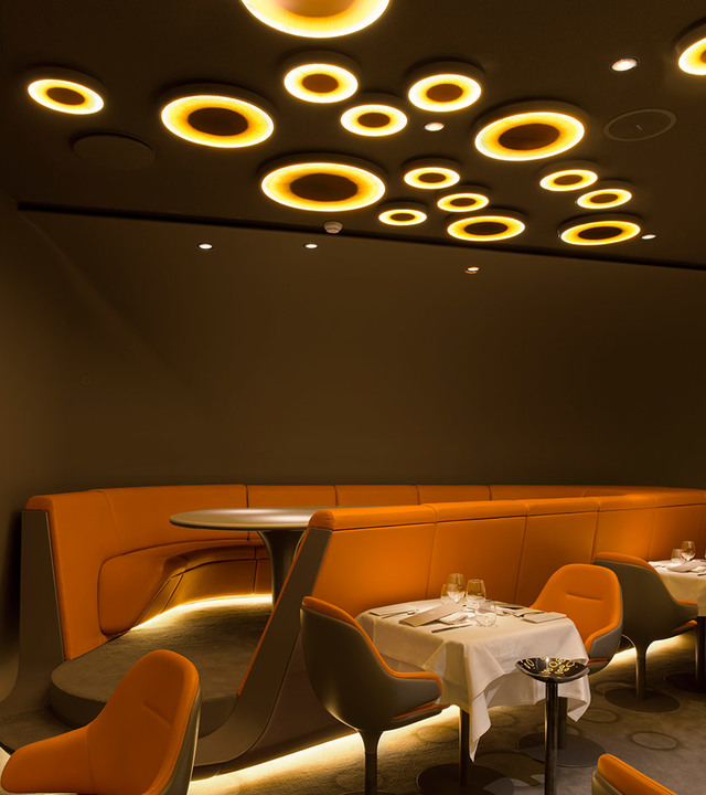 Restaurant interior with circular Artemide ceiling lights in warm gold tones above orange booth seating and white-clothed dining tables.