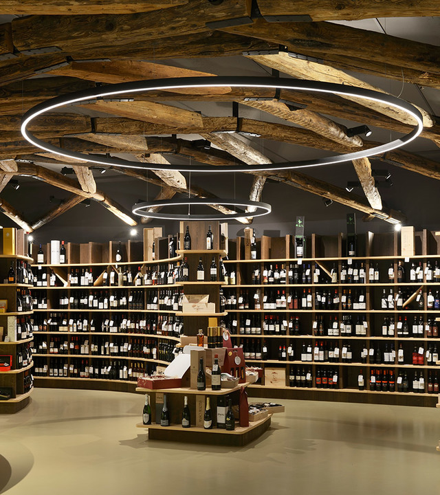 Wine shop interior with Artemide circular LED pendant rings illuminating rustic wooden beam ceiling and floor-to-ceiling wine bottle shelves