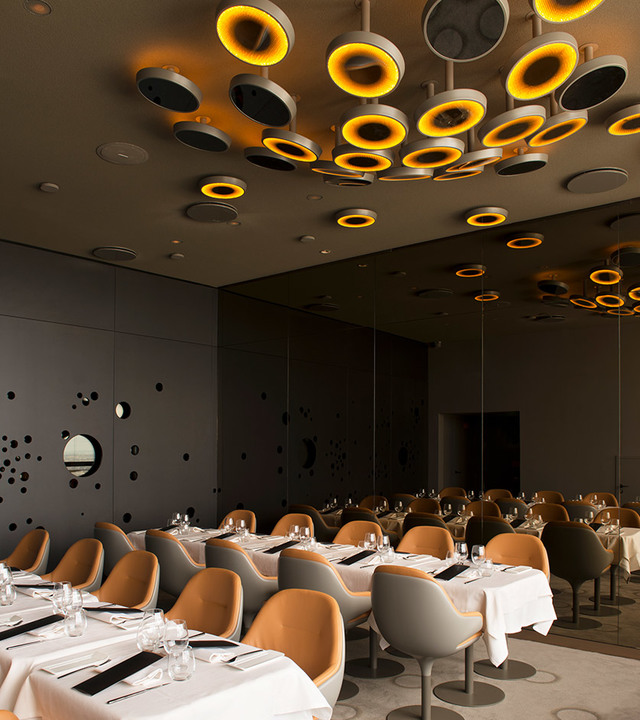 Elegant restaurant with circular Artemide pendant lights glowing amber on dark ceiling, above neatly set tables and tan upholstered chairs.